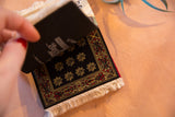 Miniature Bokhara rug coaster showing black field, yellow floral motifs, red and gold border.