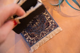 Hand lifting natural rubber backing of blue floral CoasterRug with white fringe.