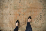 Feet on vintage distressed Oushak rug, revealing worn pile, visible foundation, faded apricot.