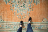 Distressed apricot orange Oushak rug with faded floral patterns and border, seen with feet.