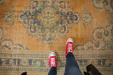Distressed Oushak rug with worn apricot field, dusty teal medallion, and visible foundation texture underfoot.
