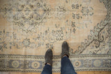 Person standing on a distressed vintage Oushak rug with faded beige field and pale blue floral motifs.