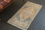 Lower corner of distressed Oushak rug with dusty blue rams head motifs, pale ochre field, and fringe.