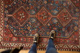 Feet on a vintage Afshar rug, highlighting sapphire blue and rust red geometric patterns.