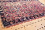 Antique Kerman rug corner, showing cochineal red field, navy floral border, and tattered edge.