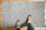 Distressed Oushak rug mat, subtle silver-grey tones with textured low pile, seen with slippers.