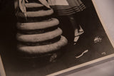 Vintage black and white photograph showing a foot, striped skirt, stacked rounds, and studio signature.