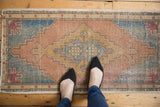 Feet on a distressed vintage Oushak rug, showcasing faded terra cotta and sky blue geometric pattern.