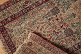 Folded Kerman rug corner reveals cochineal red border, cypress trees, and overcast side stitch.