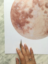 A hand holds a fine detail brush beside the Warm Moon watercolor art print.
