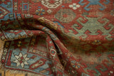 Folded antique Caucasian rug, geometric red, sky blue, and oxidized green patterns with tattered fringe.