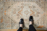 Distressed vintage Oushak rug with diamond motifs and tangerine accents under black loafers.