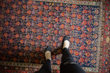 Vintage Malayer rug's germinating floral pattern in navy, cherry red, saffron, viewed with feet.