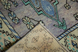 Distressed lavender and blue Oushak rug pile folded, revealing hand-knotted reverse and a corner stain.