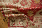 Folded distressed Oushak rug showing pale raspberry red, beige tones, and stylized animal motifs.