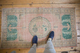 Distressed vintage Oushak rug corner with green laurel motif, pink field, and even wear showing foundation.