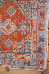 Distressed Oushak rug corner displays madder red, ivory, lavender geometric spandrel with fringe.