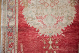 Distressed Oushak field in cherry red with serrated contour and intricate ivory border.
