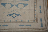 Distressed ivory Oushak rug corner showing blue geometric relief, textured pile, and brown borders.