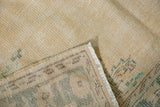 Distressed ivory Oushak carpet with pale teal and brown motifs, showing worn pile, visible foundation, and fringe.
