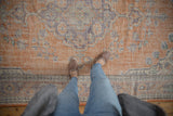 Distressed vintage Oushak rug with faded terracotta and royal blue motifs, viewed with feet.