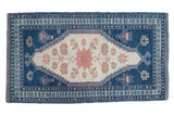 Distressed Oushak rug with central blush serrated flower, ivory field, and indigo blue patterns.