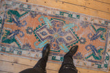 Distressed vintage Oushak runner beneath boots, showing terracotta, green, blue medallion, worn pile.