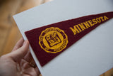 Vintage maroon felt University of Minnesota pennant with gold text and seal, held by hand.