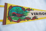 Black VERMONT text with retro reddish-orange pine cones on yellow felt pennant.