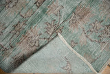 Corner of distressed Oushak rug showing jade green pile, visible foundation, and fringe.