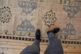 View of a distressed Oushak runner's faded geometric pattern with human feet, showcasing worn pile and beige tones.