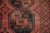 Vintage Ersari carpet close-up, dusty blush and charcoal navy geometric gul motifs, with shoes.