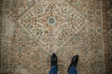 Distressed Hamadan carpet's worn texture and Herati pattern in soft tones, with boots for scale.