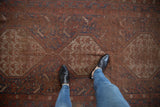 Person's feet on antique Qashqai rug, showing worn madder red pile, visible foundation.