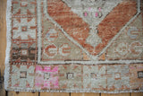Distressed Oushak rug corner with fringe, showcasing pale rust, beige, and hot pink geometric patterns.