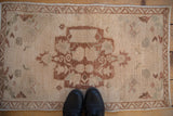 Vintage distressed Oushak mat displaying central medallion, muted floral pattern, with shoes for scale.