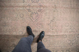 Distressed Oushak rug with person's feet, showing faded pink, ivory, and silver vinery.