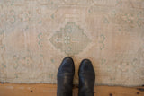 Distressed Oushak mat in cream and beige with celadon patterns, showcasing soft pile and boots for scale.