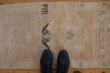 Vintage Oushak rug mat with distressed beige texture and faded seafoam motifs, with shoes for scale.