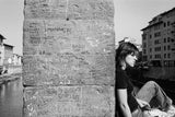 A black and white photograph of a woman sitting pensively against a stone wall covered in carved graffiti.