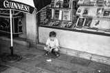 Black and white portrait of The Boy by a shop window with books and Guinness umbrella.