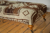 Corner of vintage Oushak rug ottoman, brown and cream geometric pattern, wooden leg.
