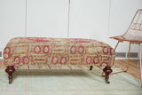 Antique rug fragment ottoman, side view: faded raspberry, saffron, ivory tribal patterns, mahogany leg.