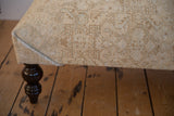 Distressed vintage Persian rug ottoman corner, showcasing muted ivory, beige tones and dark wooden leg.