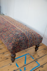 Detail of vintage Persian rug ottoman corner: faded navy, red, gold pattern, dark turned leg.