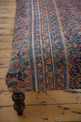 Angled detail of vintage Persian rug ottoman surface, featuring navy, faded red, gold motifs and dark wooden leg.