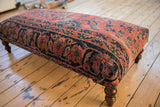 Turned wooden leg of the Persian rug ottoman on light wood floorboards.