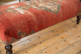 Aged red and teal geometric border of the Malayer rug fragment on ottoman, dark wooden leg.