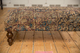 Antique Kerman rug ottoman detail, featuring worn beige, blue, red patterns and a carved wooden leg.