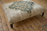 Dark curved wooden leg and textured beige fabric on the antique rug ottoman.
